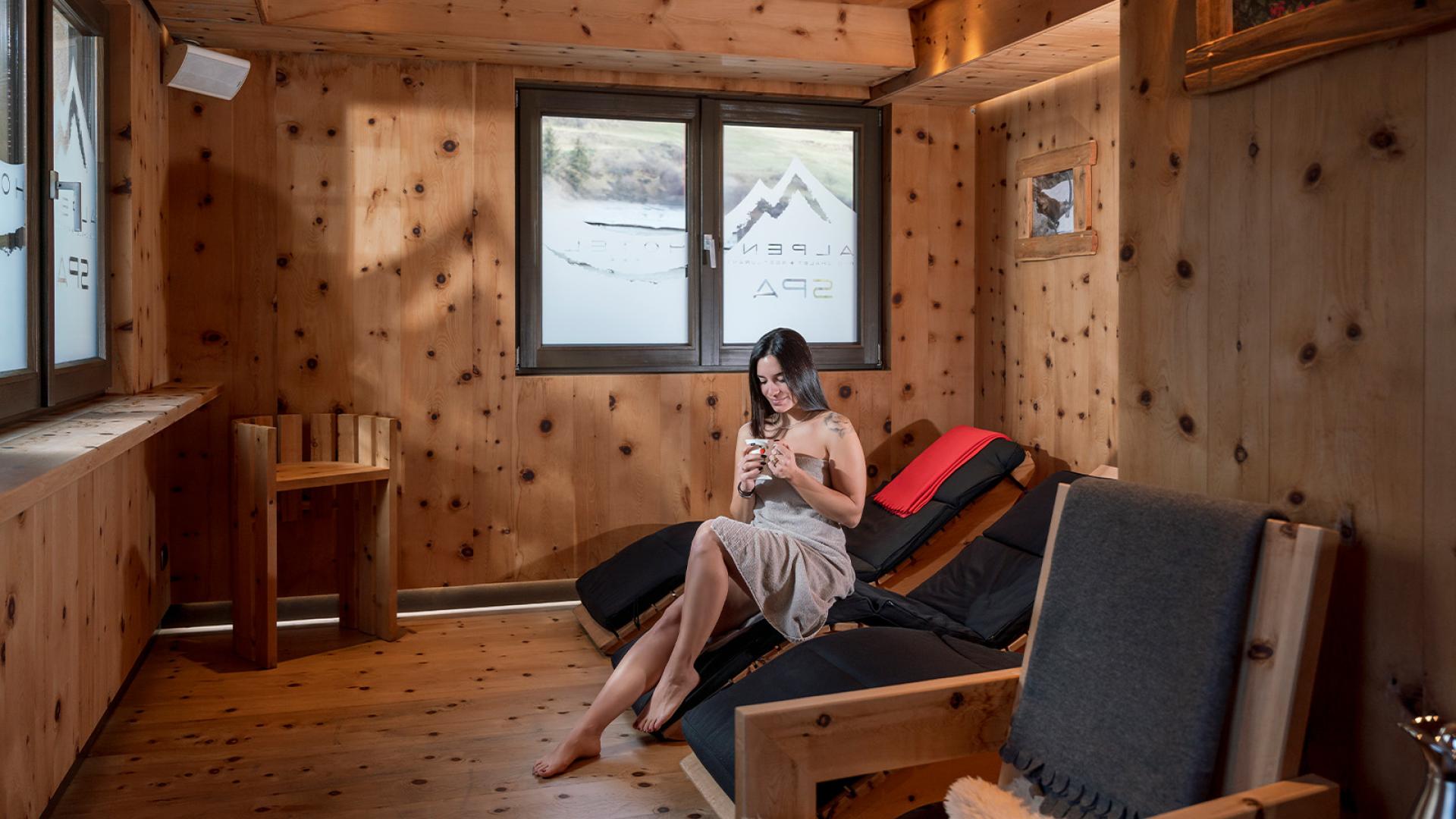 Donna rilassata in una spa in legno, seduta su una poltrona con tazza.